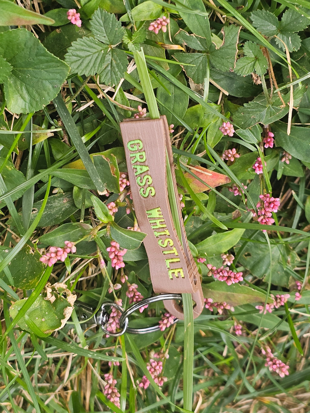 Tan grass whistle keychain with lime GRASS WHISTLE letters lying in grass and pink wildflowers