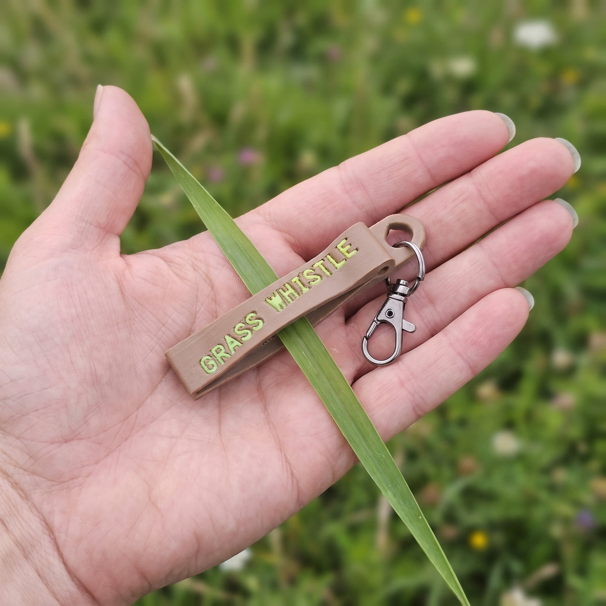 Tan grass whistle keychain held in an open hand next to a broad blade of grass