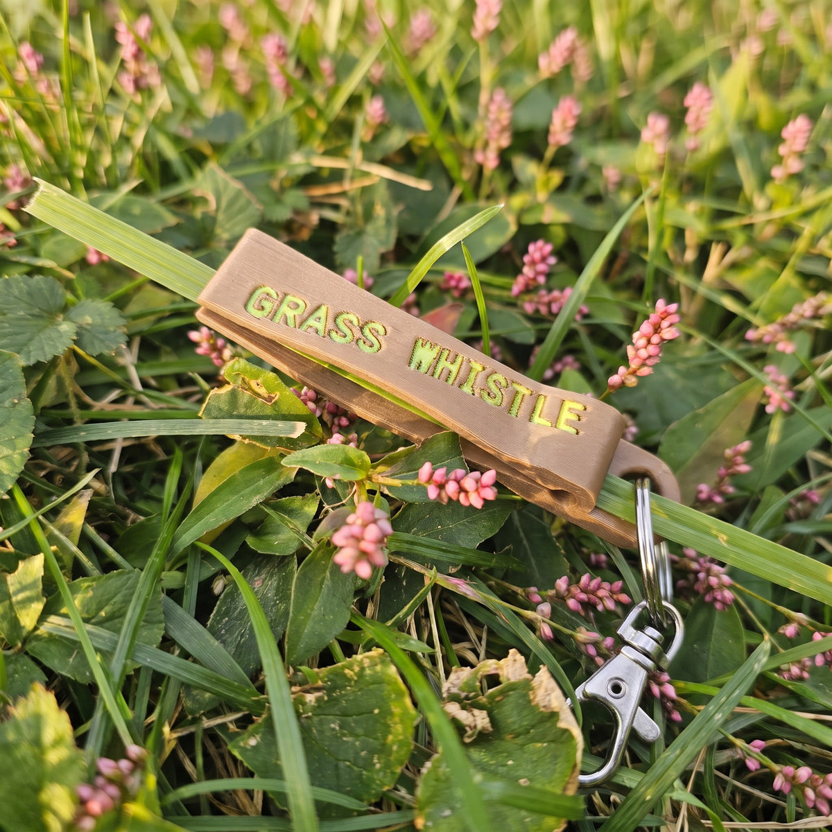 Tan grass whistle keychain with lobster clasp resting in grass